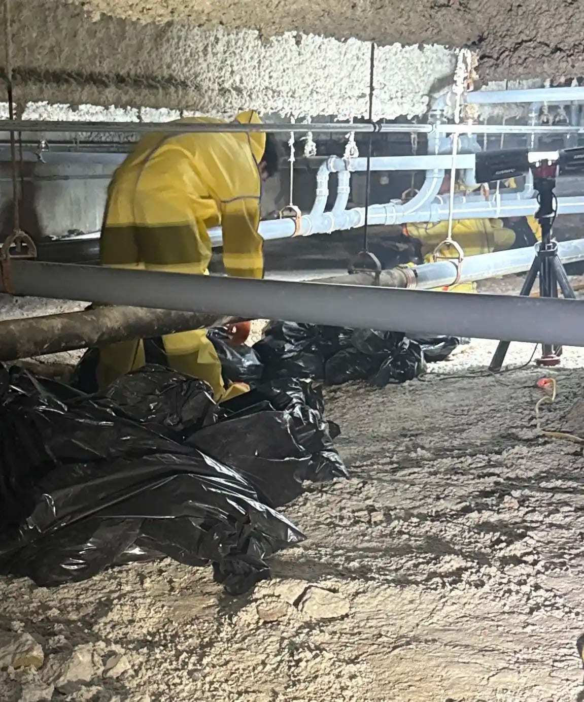 Worker in a yellow protective suit filling disposal bags in a dirt crawlspace