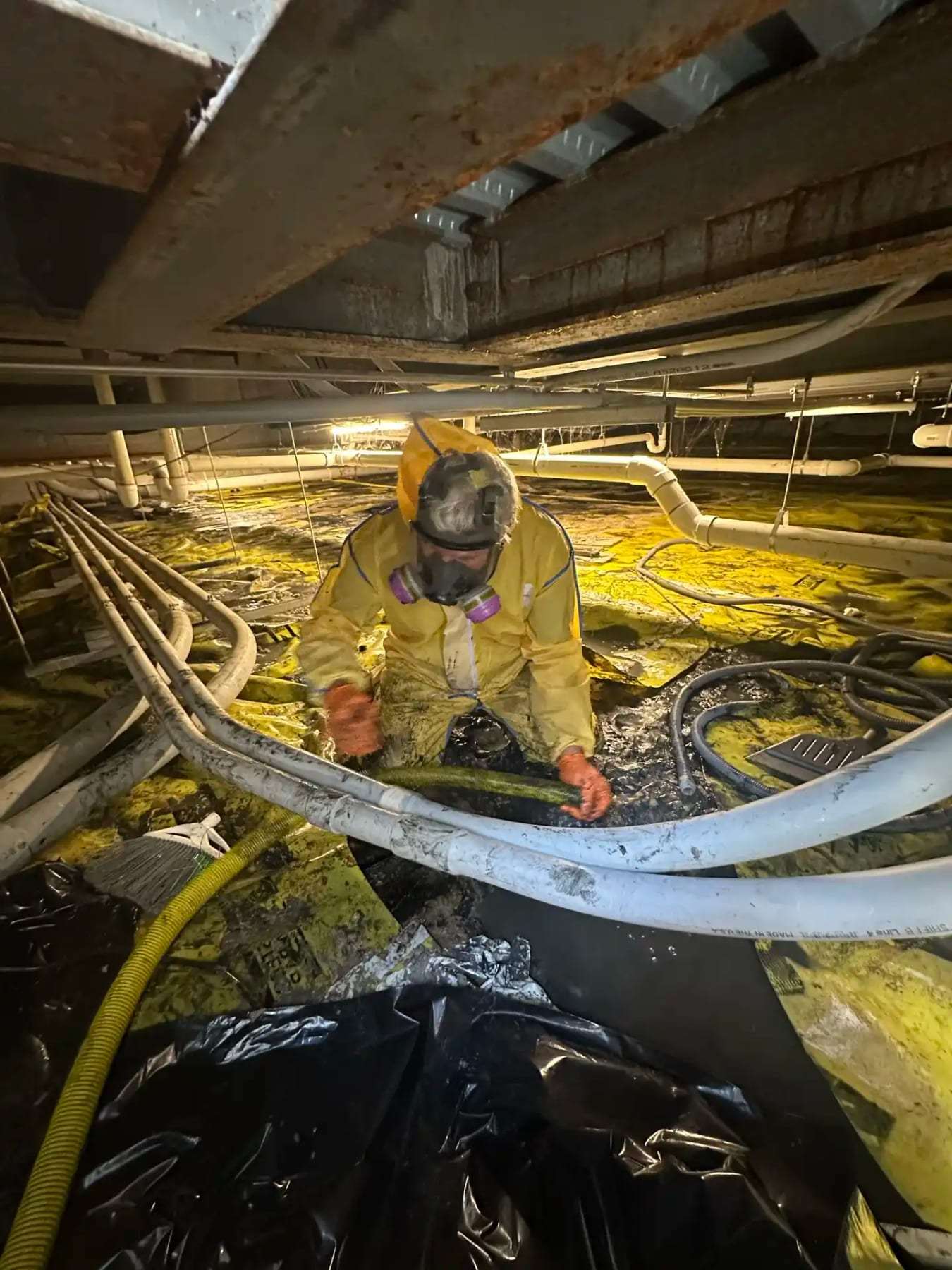 Technician in a yellow protective suit among hoses and insulation in a crawlspace